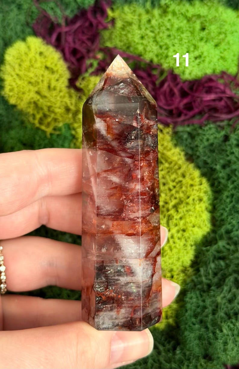 May include: A clear, rectangular crystal tower with red and white inclusions. The crystal has a pointed top and is held in a hand. The number "11" is visible on the crystal. The background is green and purple.