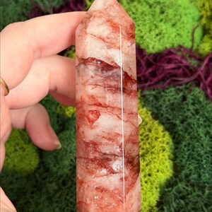 May include: A clear quartz crystal tower with red inclusions and a polished finish. The crystal is held in a hand against a backdrop of green and purple moss. The number "5" is visible on the crystal.