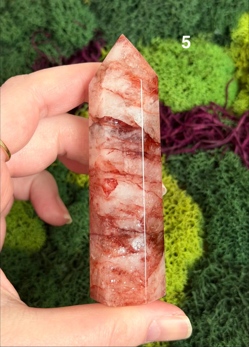 May include: A clear quartz crystal tower with red inclusions and a polished finish. The crystal is held in a hand against a backdrop of green and purple moss. The number "5" is visible on the crystal.