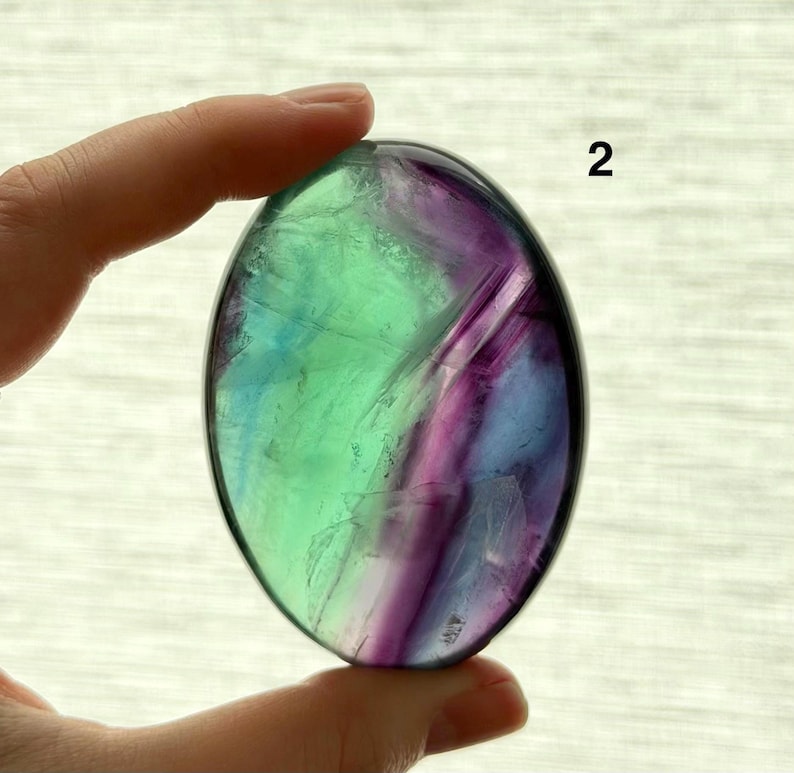 Puede incluir: Una piedra de fluorita ovalada con una superficie pulida. La piedra muestra una mezcla de colores, incluyendo tonos verdes, morados y azules. La piedra es sostenida por una mano.