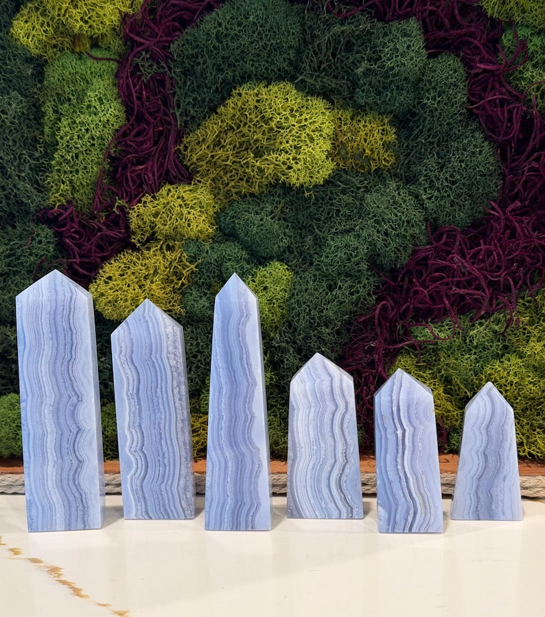 May include: Six blue lace agate obelisks with a layered, wavy pattern. The obelisks are arranged in a row, varying in height. The background features a textured wall of green, yellow, and burgundy moss.