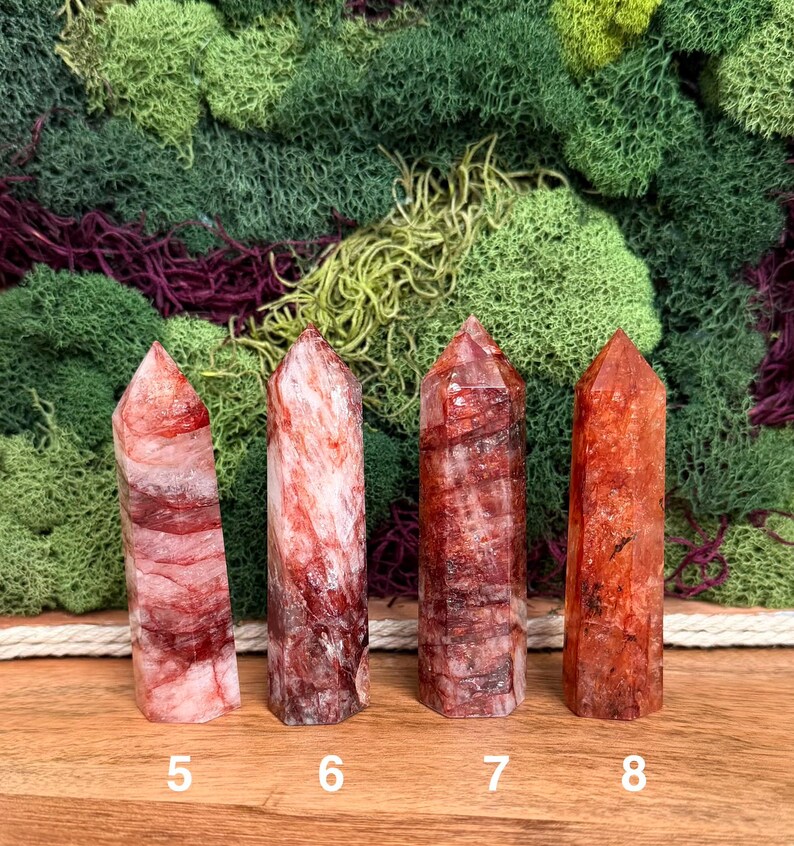 May include: Four red quartz crystal points with varying patterns and shades of red and white. The crystals are numbered 5, 6, 7, and 8. The background features green and purple moss.