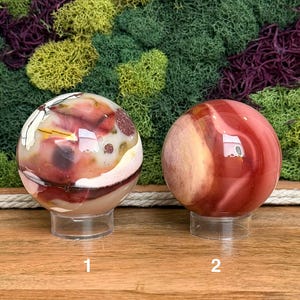Puede incluir: Dos piedras esf&eacute;ricas pulidas sobre soportes circulares transparentes. La esfera izquierda presenta patrones de remolinos en crema, rojo intenso y marr&oacute;n con peque&ntilde;as inclusiones oscuras. La derecha muestra bandas lisas de color rojo y canela.