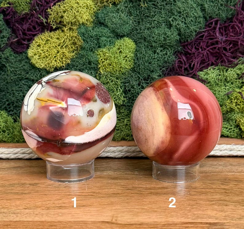 Puede incluir: Dos piedras esf&eacute;ricas pulidas sobre soportes circulares transparentes. La esfera izquierda presenta patrones de remolinos en crema, rojo intenso y marr&oacute;n con peque&ntilde;as inclusiones oscuras. La derecha muestra bandas lisas de color rojo y canela.