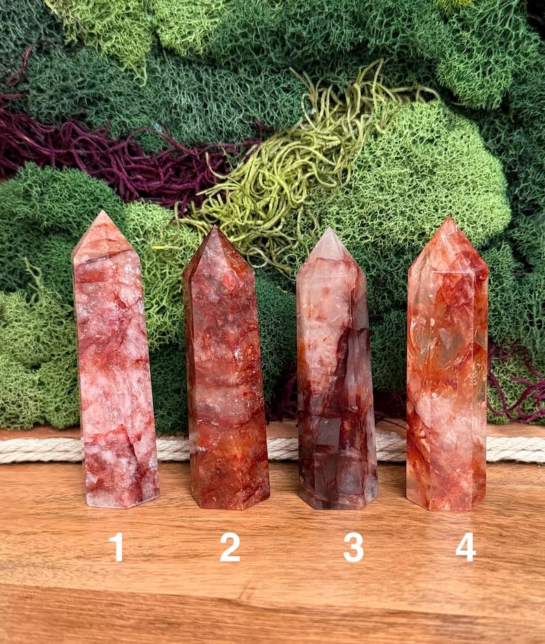 May include: Four red quartz crystal points with varying shades of red and white. The crystals are numbered 1 through 4 and are displayed on a wooden surface against a backdrop of green and burgundy moss.