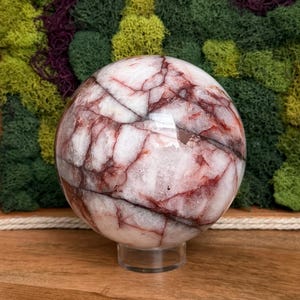 May include: A polished sphere of white and red marble, resting on a clear acrylic stand. The stone features intricate red veining throughout its surface. The sphere is displayed against a backdrop of green and purple moss.