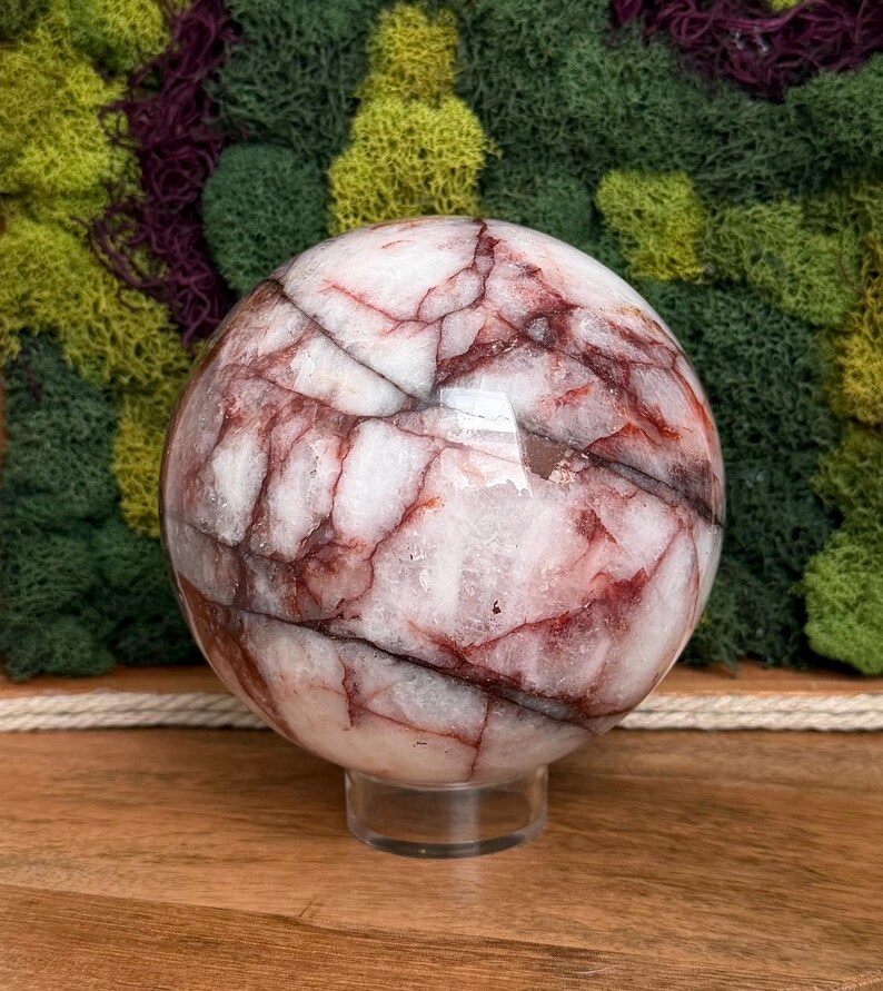 May include: A polished sphere of white and red marble, resting on a clear acrylic stand. The stone features intricate red veining throughout its surface. The sphere is displayed against a backdrop of green and purple moss.