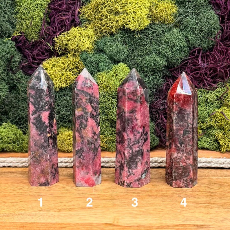 Puede incluir: Cuatro torres de cristal de rodonita pulidas con diferentes tonos de rosa, negro y gris. Cada torre tiene una parte superior puntiaguda y una base rectangular. Los cristales se exhiben sobre un fondo de musgo verde y morado.
