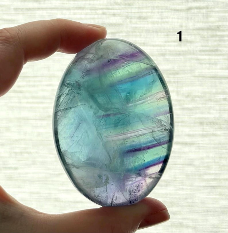 Puede incluir: Piedra de palma de fluorita ovalada con superficie pulida. La piedra presenta bandas de tonos azules, verdes y morados. El n&uacute;mero "1" es visible en la esquina superior derecha. La piedra est&aacute; sujeta por una mano.