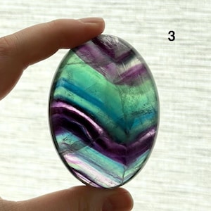 Puede incluir: Una piedra de fluorita ovalada con una superficie pulida. La piedra presenta una mezcla de colores, incluyendo p&uacute;rpura, verde y azul, con patrones de bandas visibles. El n&uacute;mero "3" es visible en la esquina superior derecha.