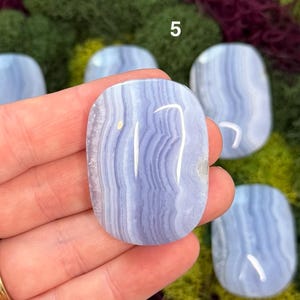 May include: A hand holds a polished, oval-shaped blue lace agate stone. The stone has a banded pattern of light and dark blue, with a white streak. The number "5" is visible in the background. Other similar stones are also visible.