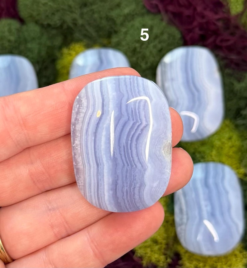 May include: A hand holds a polished, oval-shaped blue lace agate stone. The stone has a banded pattern of light and dark blue, with a white streak. The number "5" is visible in the background. Other similar stones are also visible.