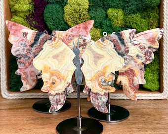 May include: Three butterfly-shaped decorative ornaments crafted from banded agate stone, featuring intricate swirls of cream, tan, brown, and pink. Each piece is mounted on a thin silver-toned metal rod attached to a circular black metal base.