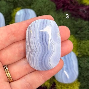 May include: A polished, oval-shaped blue lace agate gemstone with a swirling pattern of light and dark blue bands. The number "3" is visible on the top right. The gemstone is held in a hand, showcasing its natural beauty.