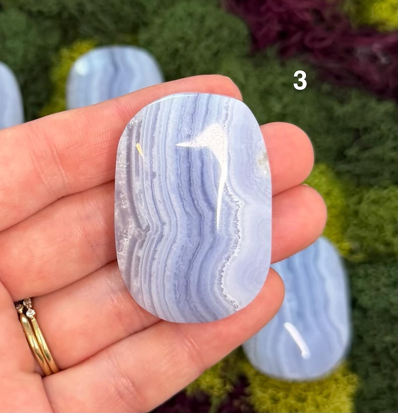 May include: A polished, oval-shaped blue lace agate gemstone with a swirling pattern of light and dark blue bands. The number "3" is visible on the top right. The gemstone is held in a hand, showcasing its natural beauty.