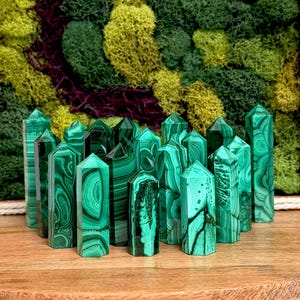 Puede incluir: Una colección de varias torres de cristal de malaquita, cada una con distintos patrones de bandas verdes y superficies pulidas, dispuestas en grupo sobre una superficie de madera con un fondo de musgo verde y morado.