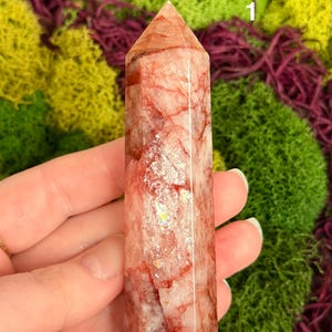 May include: A clear, rectangular crystal point with red veining and a pointed top. The crystal is held against a backdrop of green and purple moss. The crystal is approximately 4 inches tall.