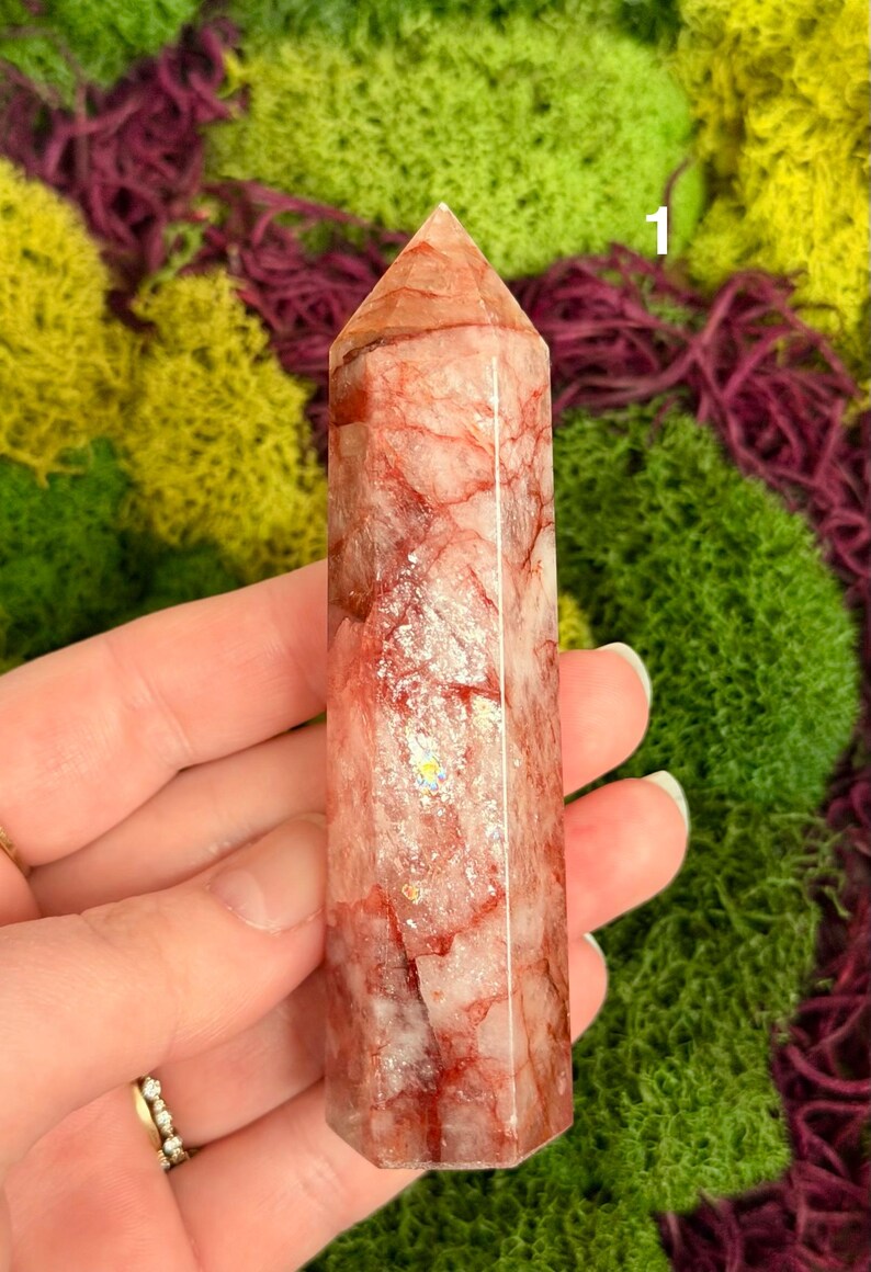May include: A clear, rectangular crystal point with red veining and a pointed top. The crystal is held against a backdrop of green and purple moss. The crystal is approximately 4 inches tall.