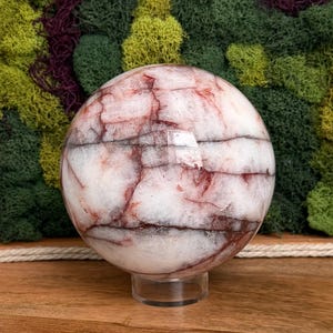 May include: A large, spherical crystal ball with a white base and intricate red and brown veining patterns. The orb is displayed on a clear, circular stand, showcasing its polished surface. The background features a mix of green and purple moss.