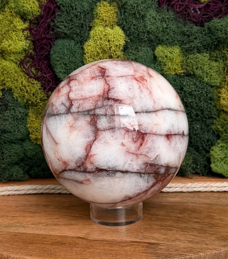 May include: A large, spherical crystal ball with a white base and intricate red and brown veining patterns. The orb is displayed on a clear, circular stand, showcasing its polished surface. The background features a mix of green and purple moss.