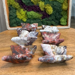 May include: A collection of eight stone sculptures, each shaped like a small boat. The sculptures display a mix of red, white, and brown colors with unique patterns. They are arranged on a wooden surface, with a decorative moss-filled frame in the background.