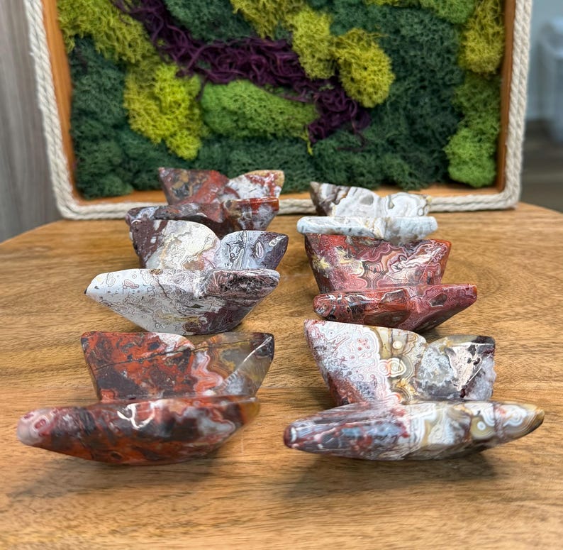 May include: A collection of eight stone sculptures, each shaped like a small boat. The sculptures display a mix of red, white, and brown colors with unique patterns. They are arranged on a wooden surface, with a decorative moss-filled frame in the background.