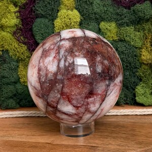 May include: A large, polished sphere of red and white stone, resting on a clear acrylic stand. The orb's surface displays intricate patterns of red veining against a white background. The sphere is approximately 4 inches in diameter.