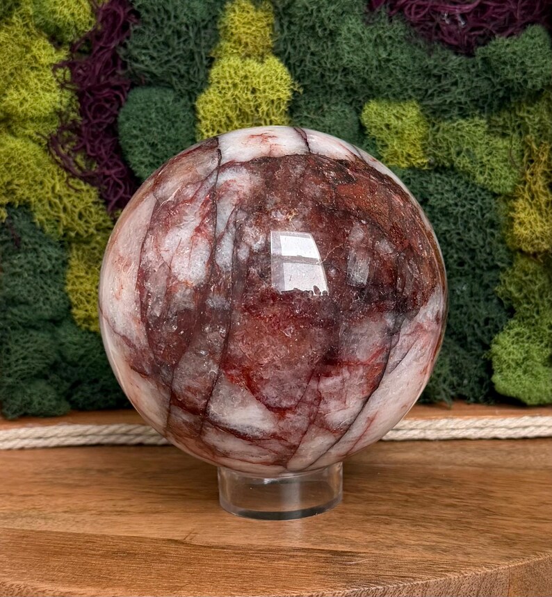 May include: A large, polished sphere of red and white stone, resting on a clear acrylic stand. The orb's surface displays intricate patterns of red veining against a white background. The sphere is approximately 4 inches in diameter.
