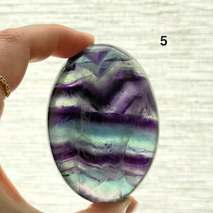 Puede incluir: Una piedra de fluorita pulida, de forma ovalada, con bandas de color p&uacute;rpura, turquesa y transparente. La piedra se sostiene en una mano, mostrando su forma lisa y redondeada. El n&uacute;mero "5" es visible en la piedra.