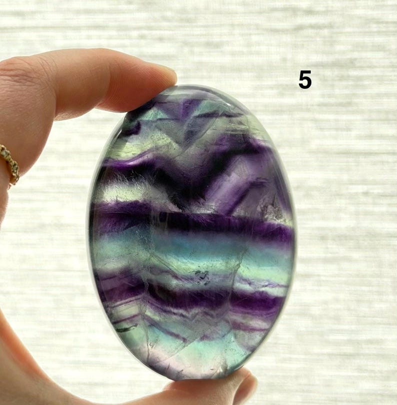 Puede incluir: Una piedra de fluorita pulida, de forma ovalada, con bandas de color p&uacute;rpura, turquesa y transparente. La piedra se sostiene en una mano, mostrando su forma lisa y redondeada. El n&uacute;mero "5" es visible en la piedra.