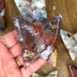 May include: A carved stone butterfly sculpture with a polished surface. The butterfly features a mix of red, brown, white, and gold colors. The sculpture is a unique decorative item.