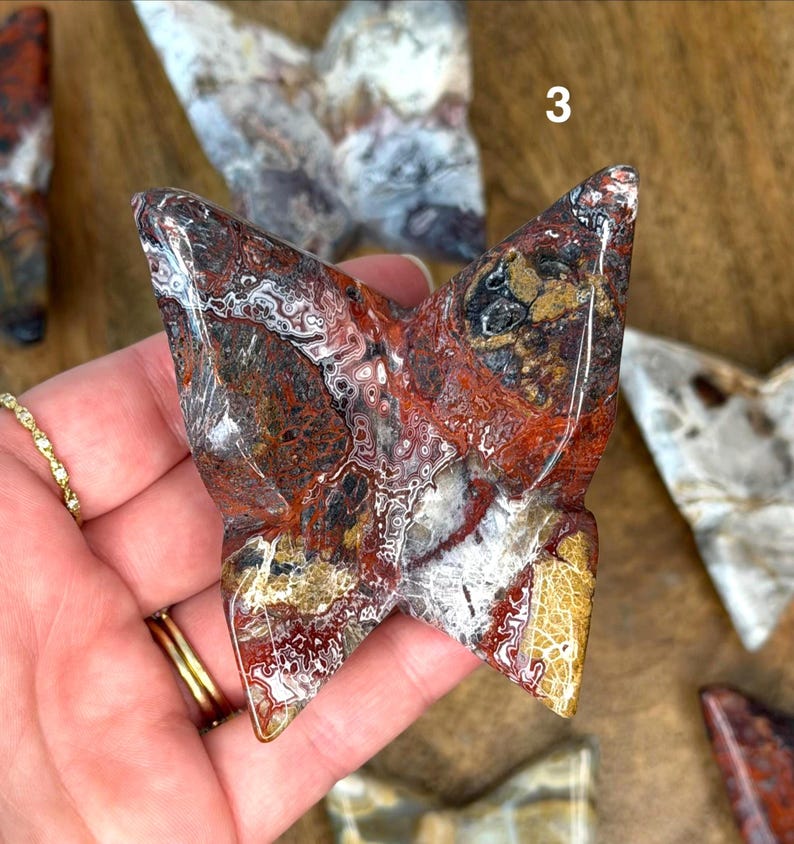 May include: A carved stone butterfly sculpture with a polished surface. The butterfly features a mix of red, brown, white, and gold colors. The sculpture is a unique decorative item.
