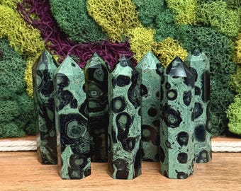Puede incluir: Una colección de siete torres de piedra pulida, cada una con un patrón distintivo de manchas circulares de color negro oscuro y verde bosque. Estas puntas de cristal hexagonales se mantienen erguidas, mostrando una superficie lisa y reflectante.