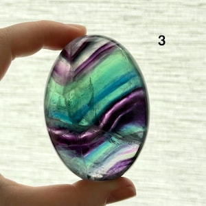 Puede incluir: Piedra de fluorita ovalada pulida, con una mezcla de colores p&uacute;rpura, verde y azul. La piedra transl&uacute;cida tiene una superficie lisa. El n&uacute;mero "3" es visible en la parte superior derecha de la piedra.