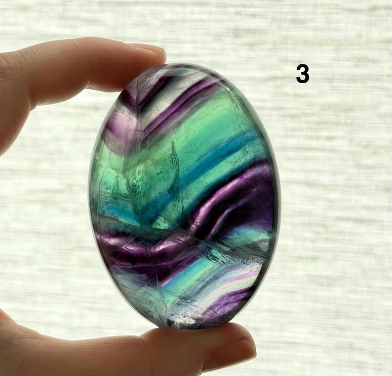 Puede incluir: Piedra de fluorita ovalada pulida, con una mezcla de colores p&uacute;rpura, verde y azul. La piedra transl&uacute;cida tiene una superficie lisa. El n&uacute;mero "3" es visible en la parte superior derecha de la piedra.