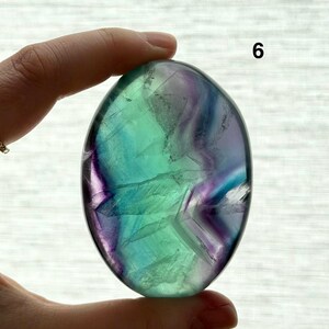 Puede incluir: Una piedra de fluorita pulida, de forma ovalada, con tonos verdes, azules y morados. La piedra est&aacute; sujeta en una mano, con el n&uacute;mero "6" visible. La superficie presenta patrones y variaciones naturales.