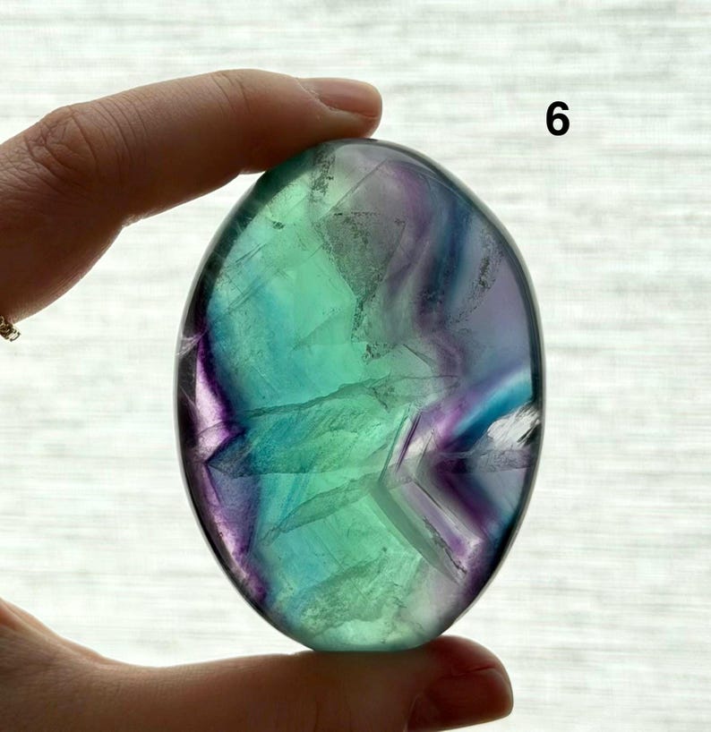 Puede incluir: Una piedra de fluorita pulida, de forma ovalada, con tonos verdes, azules y morados. La piedra est&aacute; sujeta en una mano, con el n&uacute;mero "6" visible. La superficie presenta patrones y variaciones naturales.