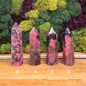 Puede incluir: Se muestran cuatro torres de piedras preciosas rosas y negras con puntas puntiagudas. Las piedras tienen un aspecto veteado con diferentes tonos de rosa y negro. Los n&uacute;meros 5, 6, 7 y 8 est&aacute;n impresos en la base de cada piedra. El fondo presenta musgos verdes, amarillos y morados.