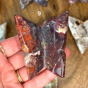 May include: A butterfly-shaped stone carving with a mix of red, black, and white patterns. The stone has a polished surface and is held in a hand. The number "9" is visible on the stone.