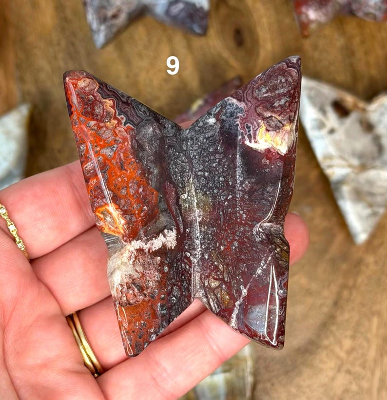 May include: A butterfly-shaped stone carving with a mix of red, black, and white patterns. The stone has a polished surface and is held in a hand. The number "9" is visible on the stone.