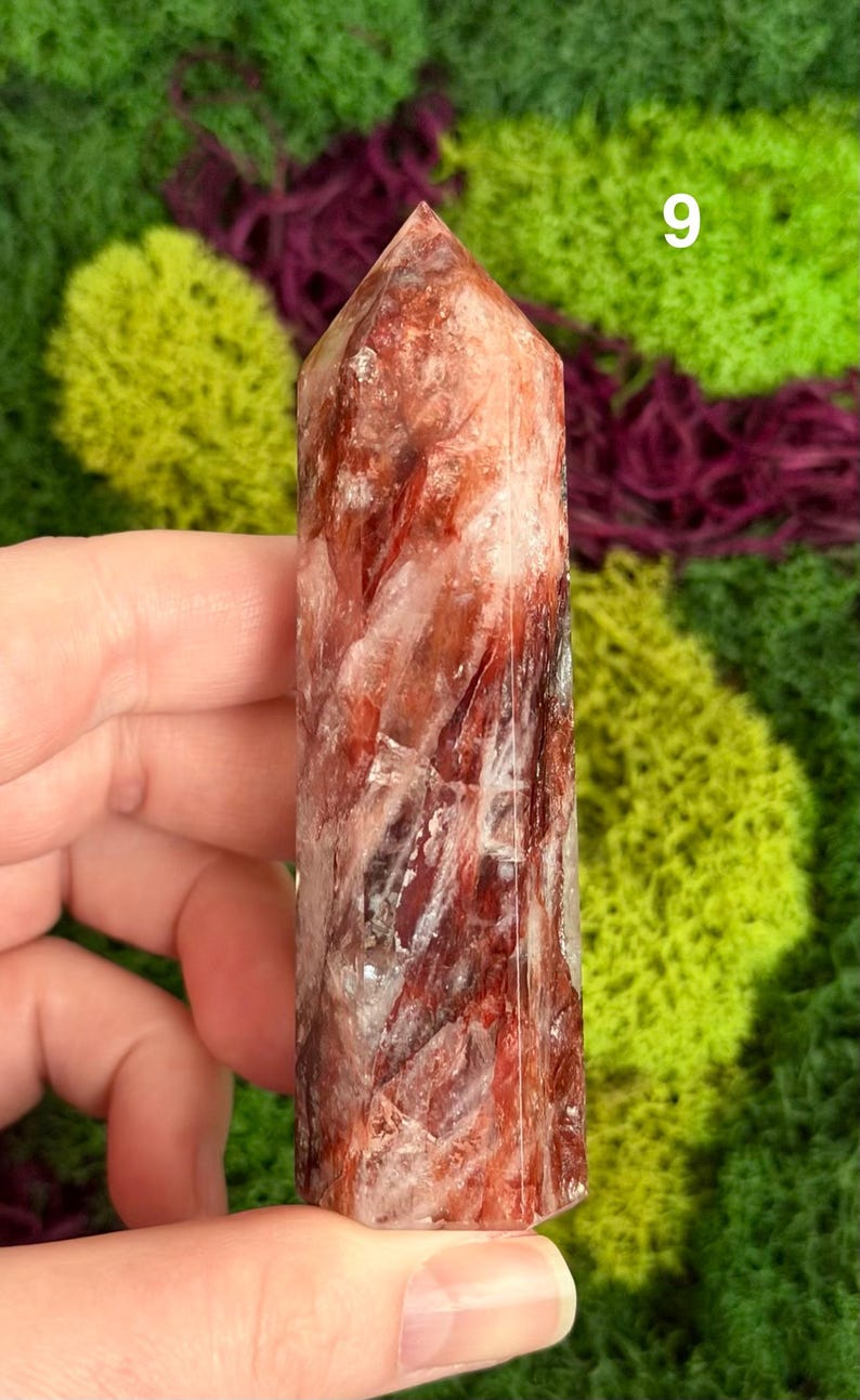May include: A red and white crystal point with a faceted top. The crystal is held in a hand against a background of green and purple moss. The number 9 is visible in the background.