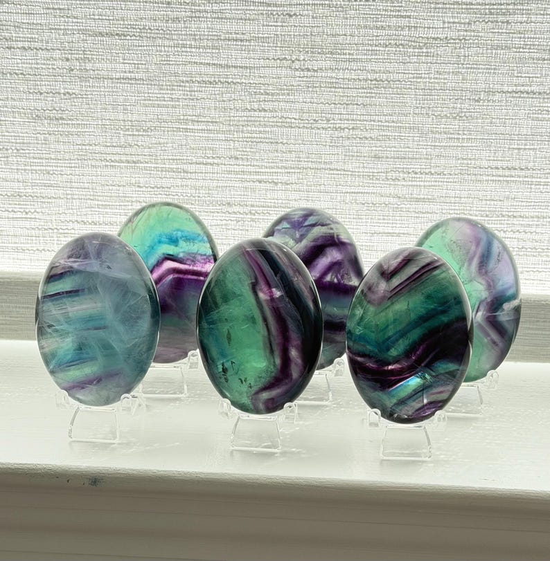 Puede incluir: Seis piedras de fluorita ovaladas en tonos morados, verdes y azules. Cada piedra se exhibe sobre un soporte acr&iacute;lico transparente. Las piedras pulidas tienen una superficie lisa y est&aacute;n dispuestas sobre una superficie blanca.