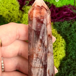 May include: A reddish-brown crystal point with a faceted surface. The crystal has a pointed top and is held in a hand. The background features green and purple moss.