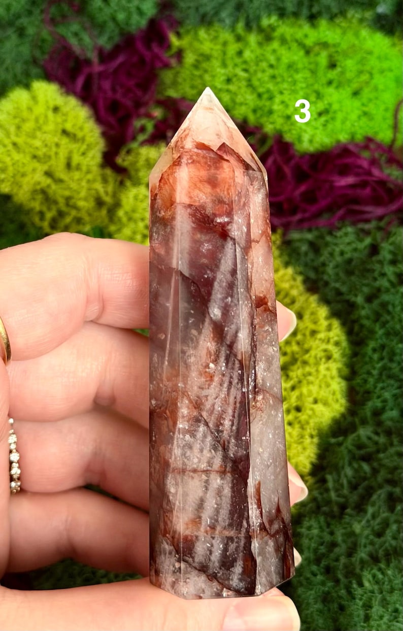 May include: A reddish-brown crystal point with a faceted surface. The crystal has a pointed top and is held in a hand. The background features green and purple moss.