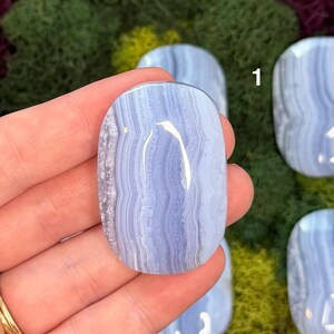 May include: A polished, oval-shaped blue lace agate gemstone. The stone features a light blue color with swirling, banded patterns. The gemstone is held in a hand, with other similar stones visible in the background.