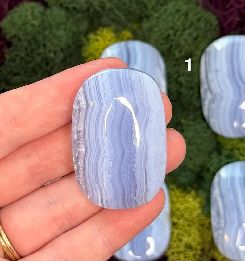 May include: A polished, oval-shaped blue lace agate gemstone. The stone features a light blue color with swirling, banded patterns. The gemstone is held in a hand, with other similar stones visible in the background.