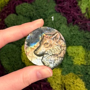 May include: A round, iridescent labradorite stone with a carved wolf head. The stone displays a range of colors, including gold, blue, and gray. The wolf is depicted in profile, with detailed fur and facial features. The stone is held in a hand.
