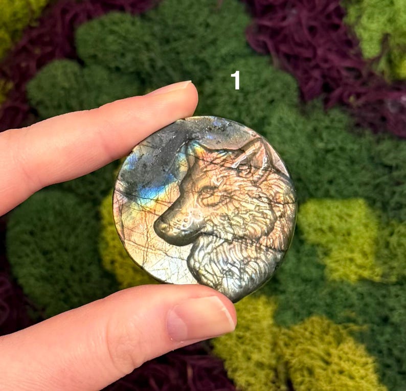 May include: A round, iridescent labradorite stone with a carved wolf head. The stone displays a range of colors, including gold, blue, and gray. The wolf is depicted in profile, with detailed fur and facial features. The stone is held in a hand.
