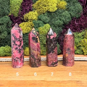 Puede incluir: Cuatro torres de cristal de rodonita pulidas, rosas y negras. Cada torre est&aacute; numerada 5, 6, 7 y 8. Los cristales se exhiben sobre una superficie de madera con un tel&oacute;n de fondo de musgo verde, amarillo y morado.