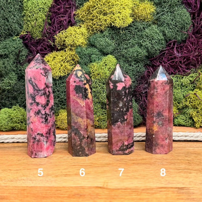 Puede incluir: Cuatro torres de cristal de rodonita pulidas, rosas y negras. Cada torre est&aacute; numerada 5, 6, 7 y 8. Los cristales se exhiben sobre una superficie de madera con un tel&oacute;n de fondo de musgo verde, amarillo y morado.
